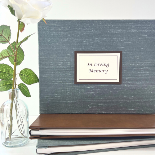 Blue silver silk cover memorial guest book shown stacked on brown leather guest book with flower prop